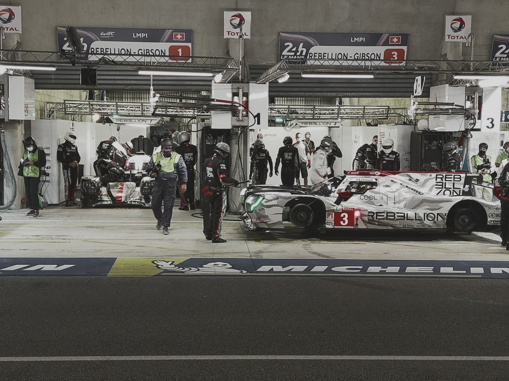 Rebellion LMP1 car in the pits during the 24 Hours of Le Mans, surrounded by mechanics during a night stop.